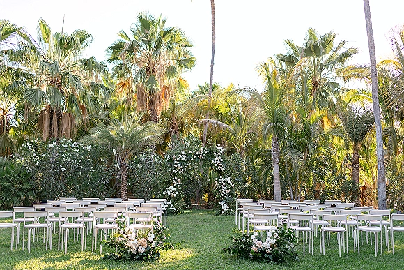 Ceremony setup for an outdoor wedding ceremony with white chairs flanking a grassy aisle lined with blush blooms and greenery, palm garden backdrop
