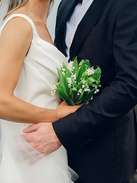 Couple portrait of bride and groom close up, embracing with a lily of the valley bouquet, her veil over a white dress against a neutral wall