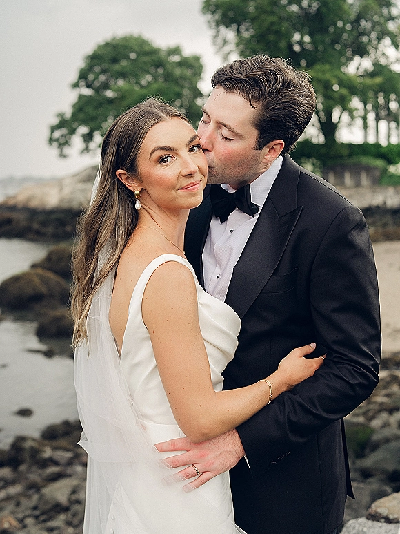 Wedding couple portrait with groom kissing bride’s cheek as she looks at camera, long veil over satin dress by rocky shoreline water