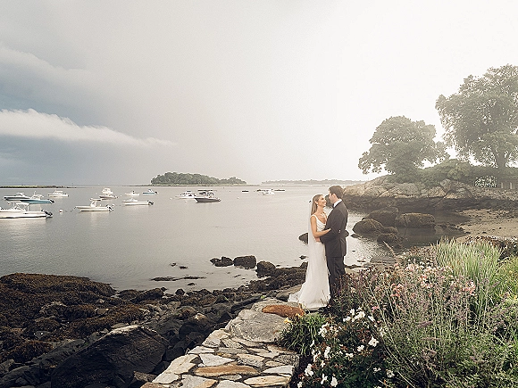 Couple portrait of bride and groom embracing on a rocky shoreline, her veil flowing, with ocean, boats, and cloudy sky behind