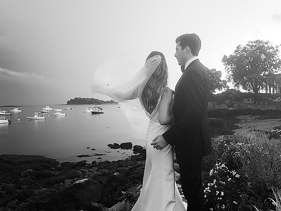 Couple portrait in black and white, bride and groom from behind with veil blowing in wind, overlooking ocean and boats by rocky shore