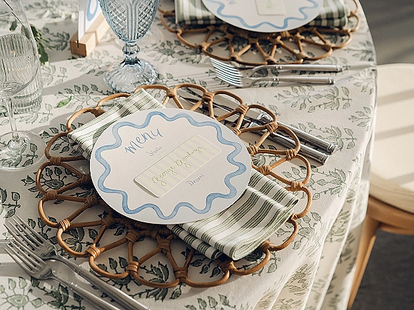 Reception tablescape with wedding place setting on a floral tablecloth, woven rattan placemat, striped napkin, menu card, and blue goblet indoors