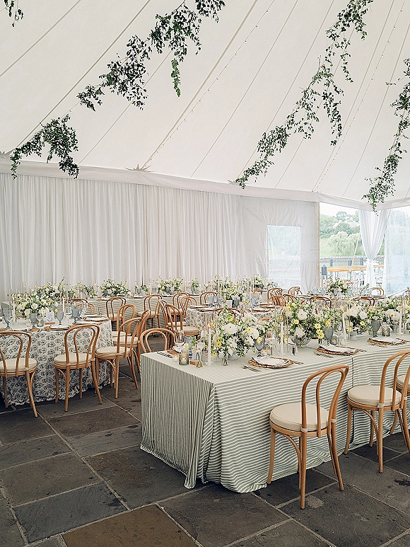 Reception tablescape with wedding banquet tables in striped linens, white florals, taper candles, and overhead greenery under a draped tent