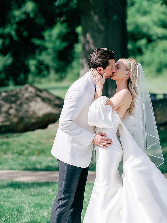 Wedding kiss portrait of bride and groom kissing, veil flowing as she cups his face, on a green lawn by trees and stone wall