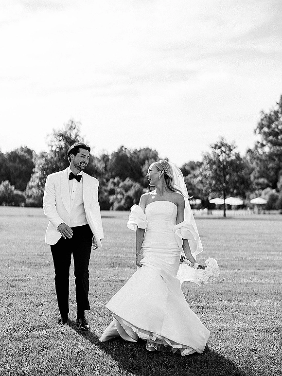 Couple portrait of bride and groom walking, bride looking at groom, holding a bouquet on a grassy lawn with trees and sky behind