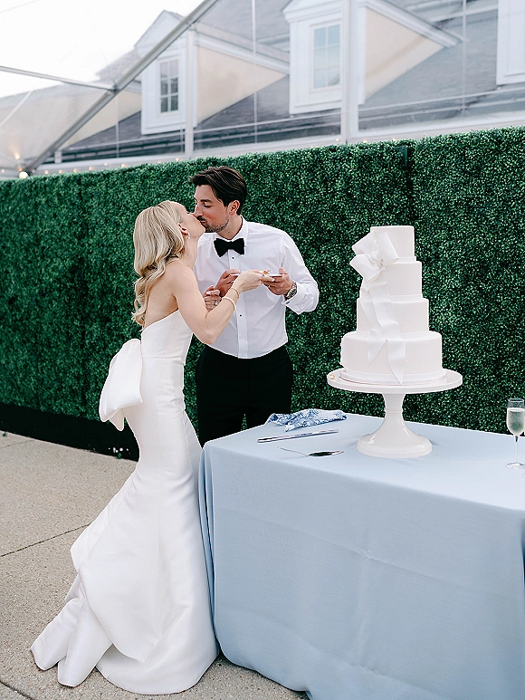 Cake cutting moment as bride and groom slice a white tiered wedding cake on a stand, champagne flute on blue tablecloth before hedge wall
