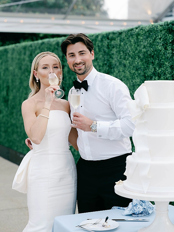 Wedding couple portrait of bride and groom champagne toast beside a white tiered cake under string lights against a green hedge wall