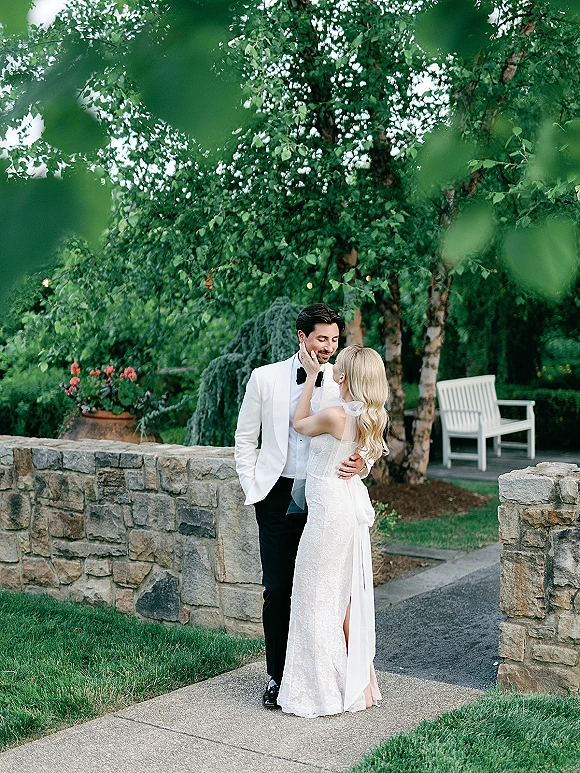 Couple portrait of a bride touching groom face, her bridal veil draped as they embrace by a stone wall on a garden walkway