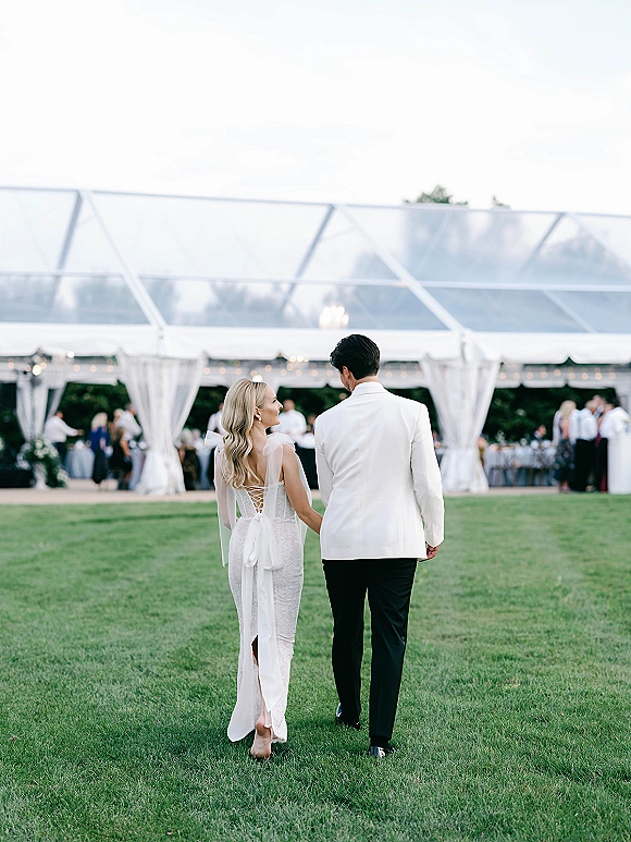 Newlywed couple holding hands walk away, bride in lace-up back gown with bow and long ribbons, under clear tent with string lights