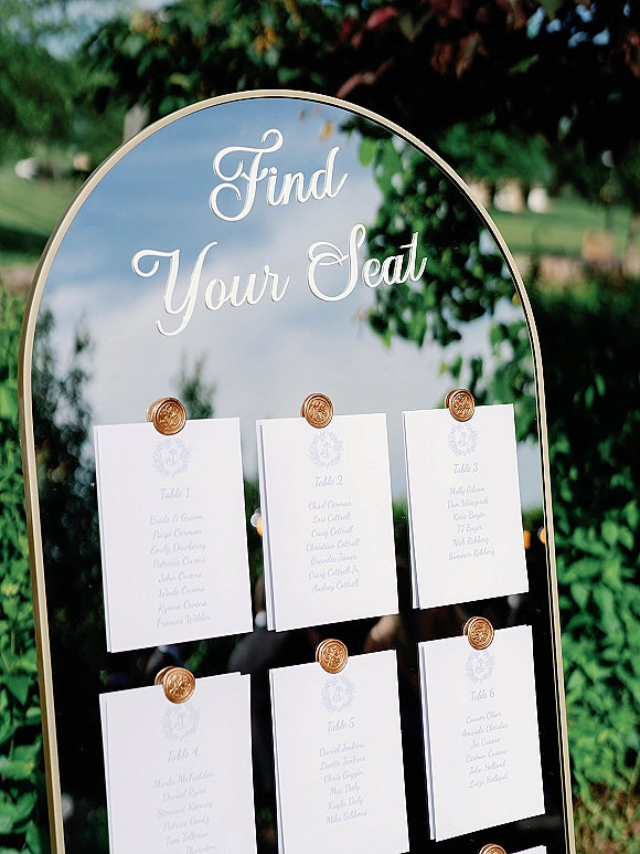 Wedding seating chart mirror seating chart on an arched gold-framed mirror with calligraphy cards and wax seals on a lawn with trees