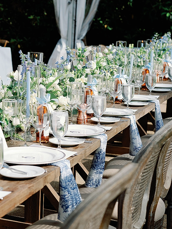 Reception tablescape with blue and white toile runner, taper candles, and floral garland on a wood banquet table on an outdoor patio