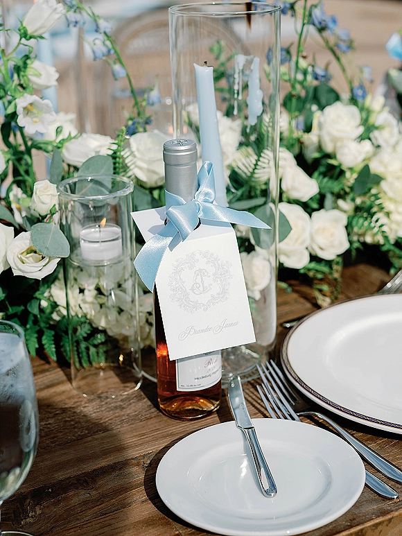 Wedding tablescape with reception table setting featuring white roses and ferns, taper candles, wine bottle favor with blue ribbon on rustic wood table