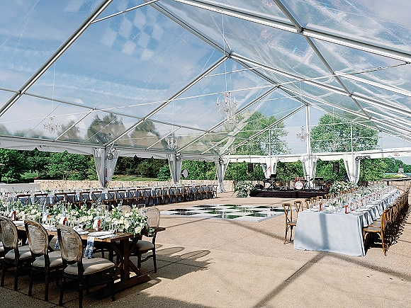 Wedding reception tent with a clear top, white draping, and crystal chandeliers over long banquet tables and checkered dance floor on a lawn