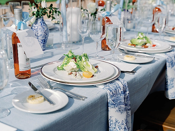 Reception tablescape with a blue and white wedding tablescape runner, light blue linen, taper candles, greenery vase, and salad place settings outdoors