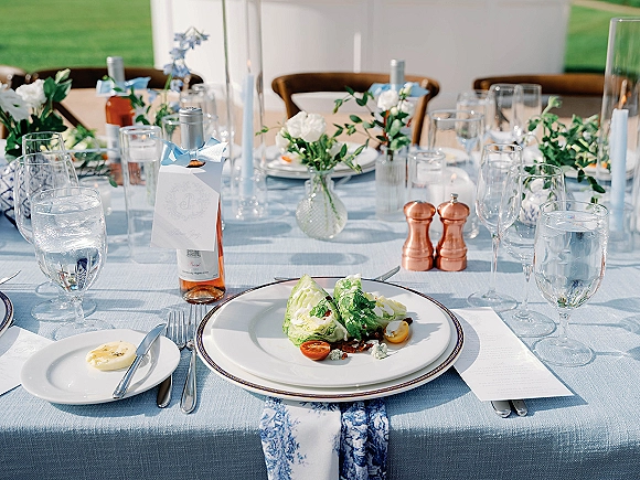 Reception tablescape with blue wedding tablescape details, light blue cloth, blue-rimmed plates, menus, taper candles, florals, rosé bottles on a lawn