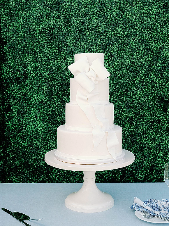 Wedding cake with a tiered white design and fondant bow, cascading ribbon on a stand with server and forks, set before a green hedge wall