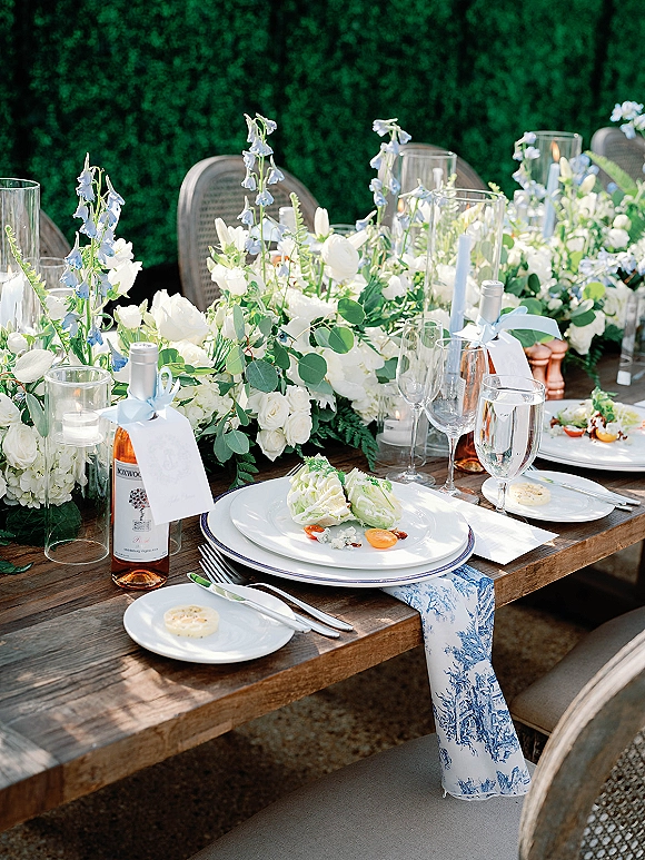Reception tablescape with outdoor wedding tablescape details: white and blue florals, greenery garland, taper candles, and tagged wine bottles by a green hedge