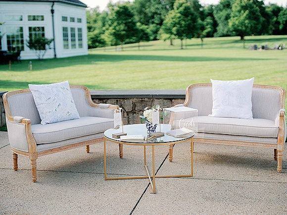 Wedding lounge seating with upholstered settees and pillows around a round glass coffee table, plus guestbook and acrylic card box on patio