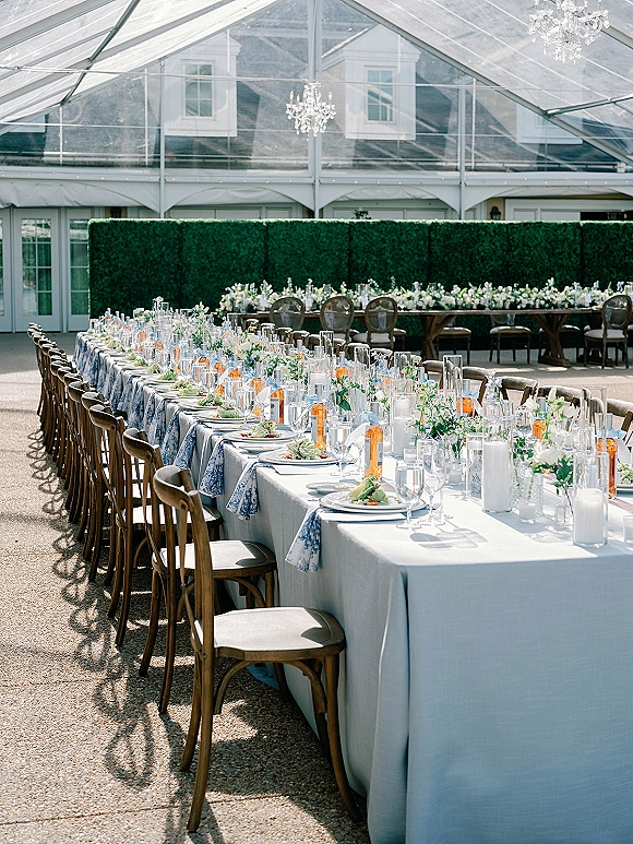 Reception tablescape with a long banquet table setup featuring blue patterned runners, white florals, candles, and chandeliers under a clear tent canopy