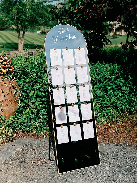 Seating chart sign on an arched mirror seating chart with gold frame, printed escort cards and wax seals on an easel in a garden path setting