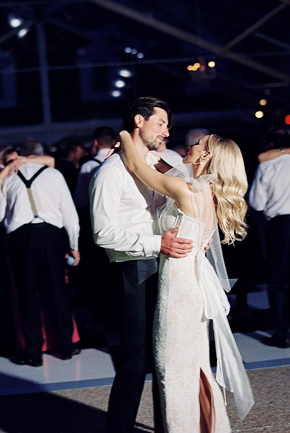 First dance as bride in a veil and strapless lace wedding dress slow dances with groom in white shirt and suspenders under string lights