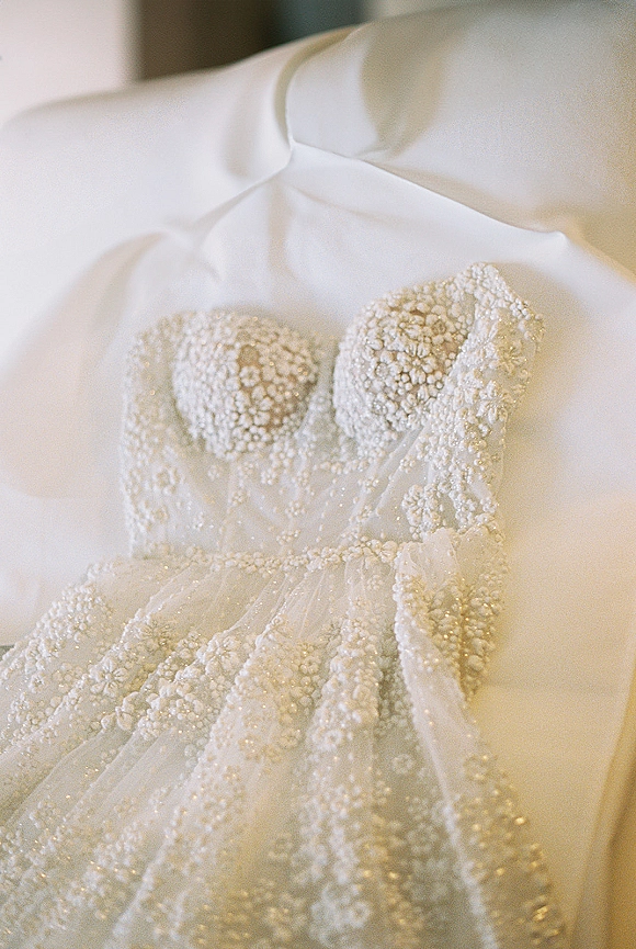 Wedding dress with a beaded wedding dress bodice featuring lace appliques and illusion tulle straps, hanging against soft neutral fabric indoors