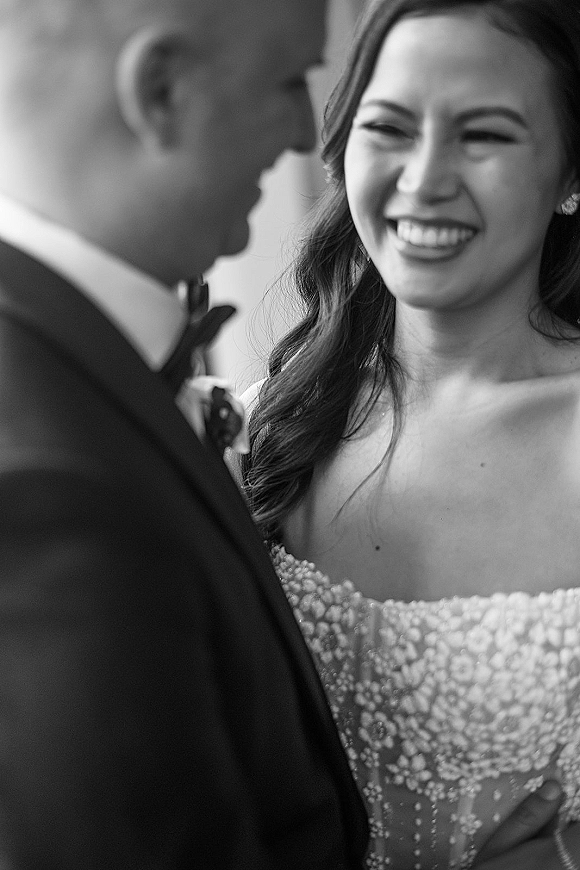Wedding couple portrait in black and white as groom whispers and bride laughs, close-up in soft natural light by an indoor wall