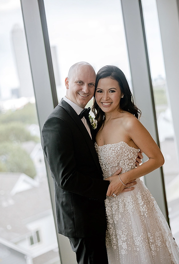Couple portrait of bride and groom smiling and hugging by floor-to-ceiling windows, her strapless wedding dress in soft natural light