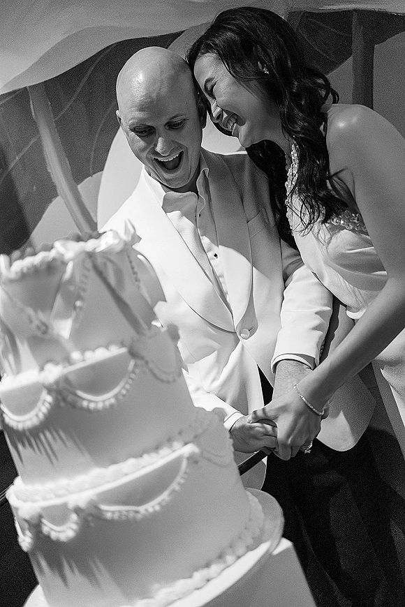 Cake cutting moment as bride and groom laugh with hands on the wedding cake knife beside a white tiered cake in soft indoor light