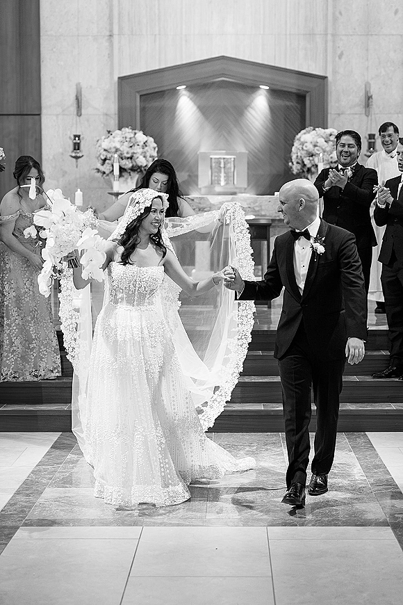 Wedding recessional as bride and groom walk the aisle holding hands, her orchid bouquet and long veil trailing past clapping guests at the church altar