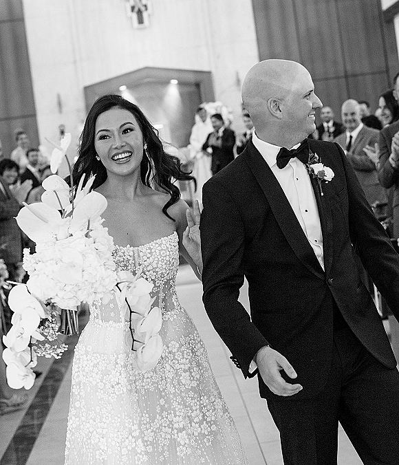 Wedding recessional as bride smiles flashing a peace sign with orchid bouquet, walking the church aisle beside groom in tuxedo as guests clap