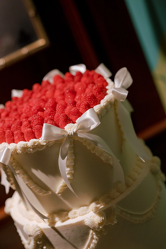 Wedding cake with raspberries on top, piped buttercream frosting and white ribbon bows, displayed indoors near dark wood furniture on green carpet