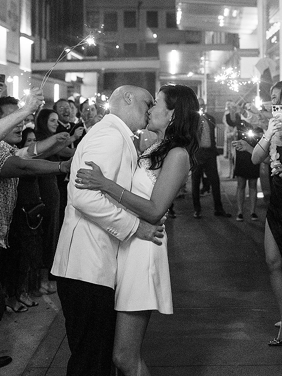 Wedding kiss during sparkler send off as guests line a night city street, bride in short dress and groom in white jacket under glowing lights