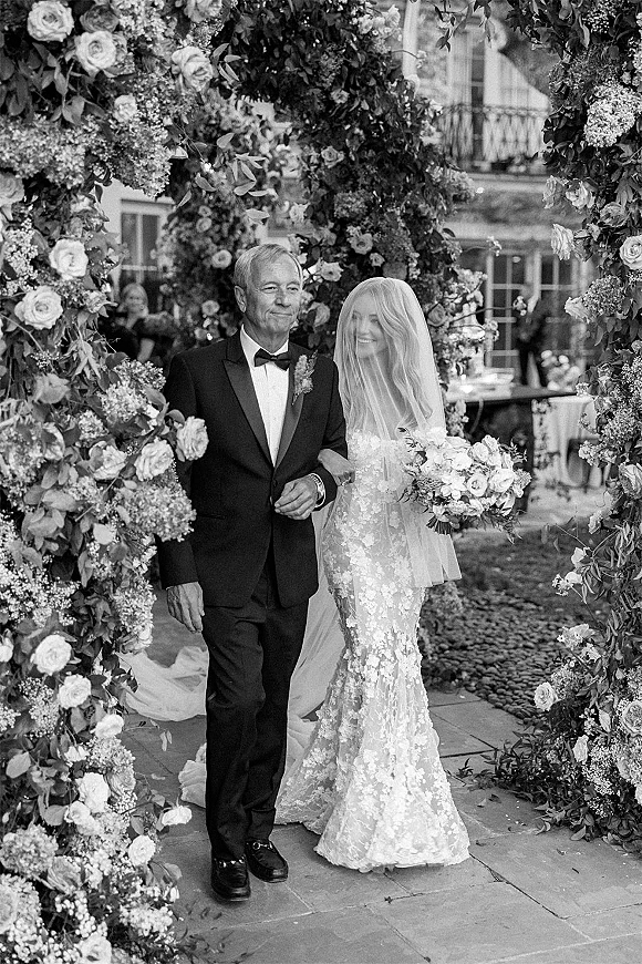 Bridal processional as bride walks down aisle with her father, cathedral veil and bouquet, entering under a lush floral arch in courtyard