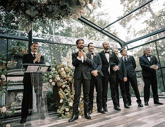 Groomsmen ceremony with groomsmen lined up in black tuxedos and boutonnieres, standing by a floral arch and clear podium in a glass greenhouse