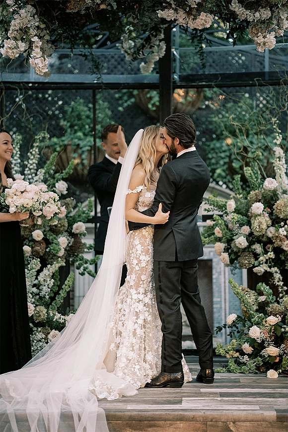 Wedding kiss under a floral arch, bridal veil flowing over a lace dress as groom in tuxedo embraces in a greenhouse with glass walls