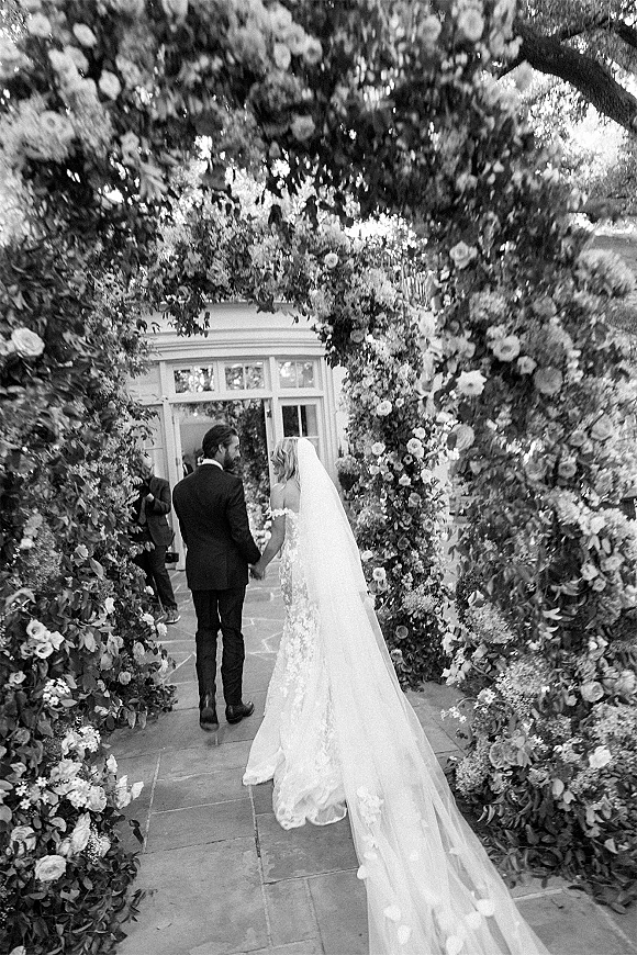 Wedding recessional bride and groom walking away holding hands under a floral arch, her long veil and train trailing on a stone garden walkway