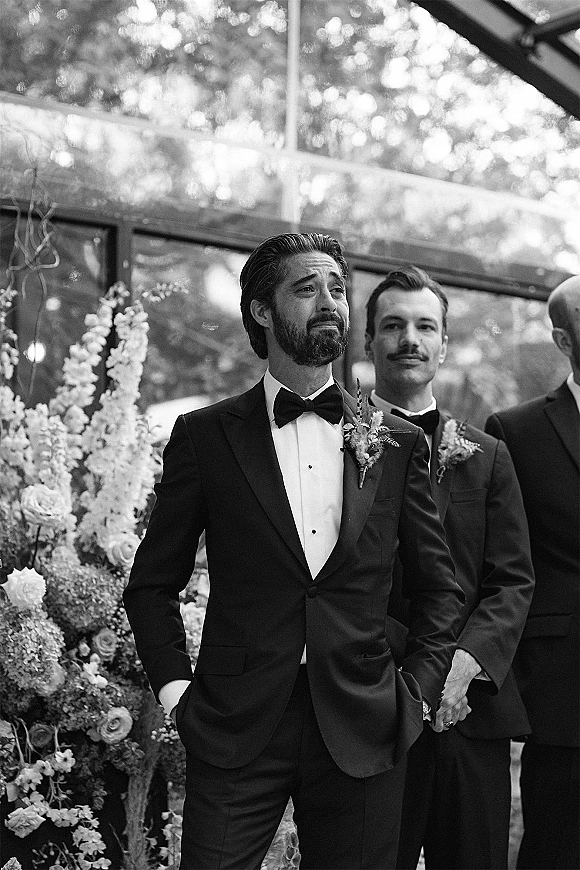 Groom portrait in a black and white wedding portrait, wearing a tuxedo and bow tie with boutonniere by a greenhouse glass wall and trees