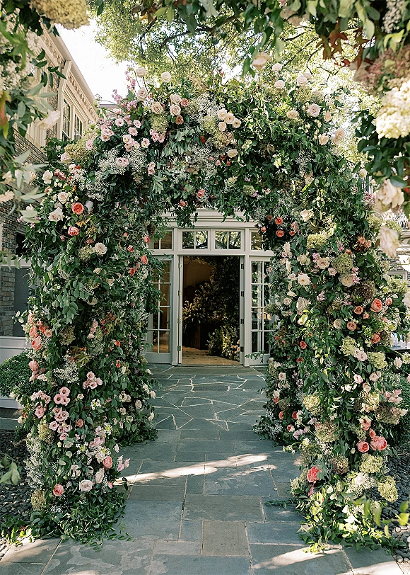 Wedding floral arch with greenery wedding arch styling, roses, hydrangea and vines framing a stone walkway by glass doors in a garden setting