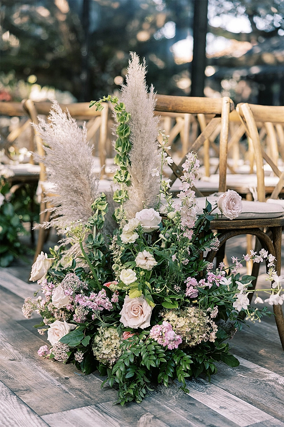 Wedding aisle flowers in a low ground floral arrangement with pampas grass, blush roses, and greenery along a wooden garden aisle with crossback chairs