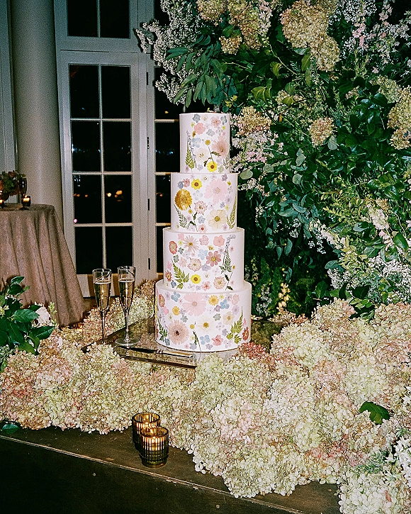 Wedding cake with pressed flower design on a linen-draped table, framed by greenery and hydrangeas with candles by glass doors