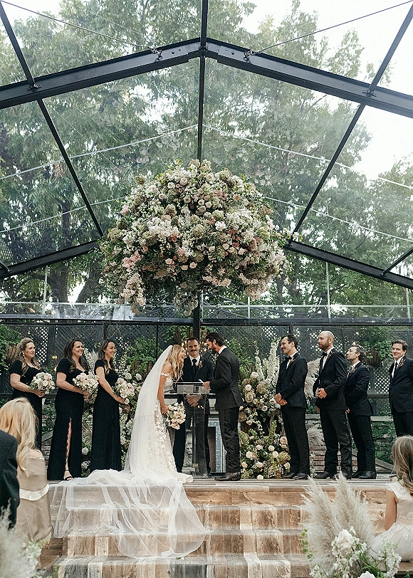 Ceremony moment at a greenhouse wedding ceremony as bride and groom stand beneath a hanging floral installation with wedding party nearby