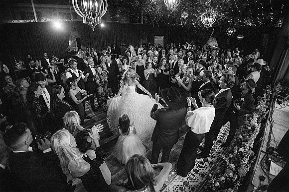 First dance on the wedding dance floor as bride in strapless ball gown and lace gloves twirls, guests in cowboy hats circle under chandeliers