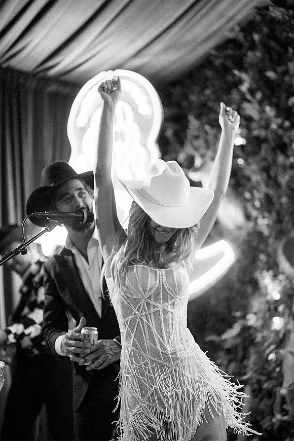 Wedding reception dancing as bride in a cowboy hat and fringed beaded mini dress raises her arms on a stage with neon sign and curtains