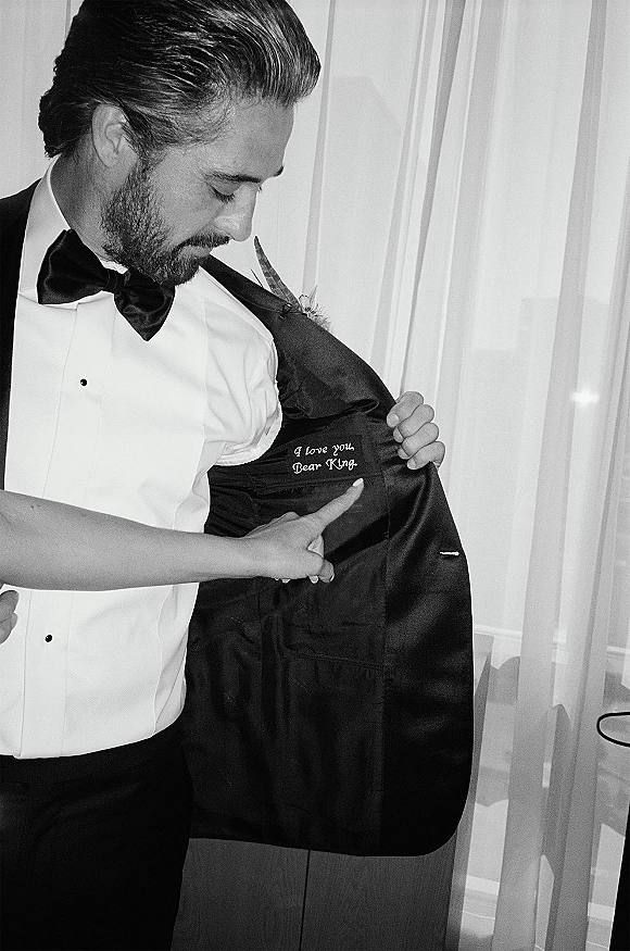 Groom getting ready in a groom tuxedo portrait, fastening his bow tie with a boutonniere, framed by curtains and an interior wall