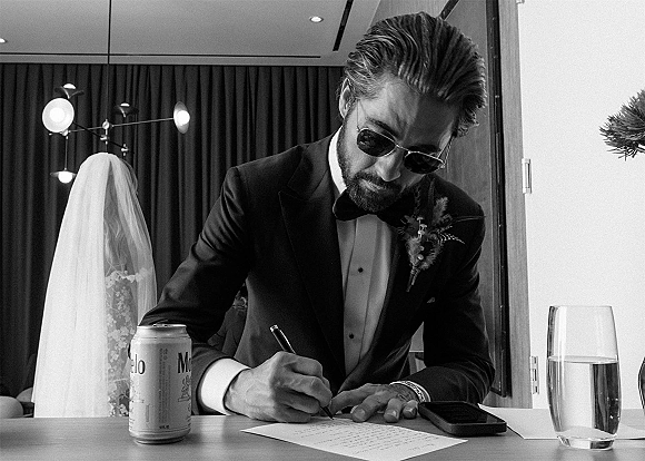 Groom getting ready, writing vows in a black tuxedo with bow tie and boutonniere beside a table, mirror, and curtain drapes
