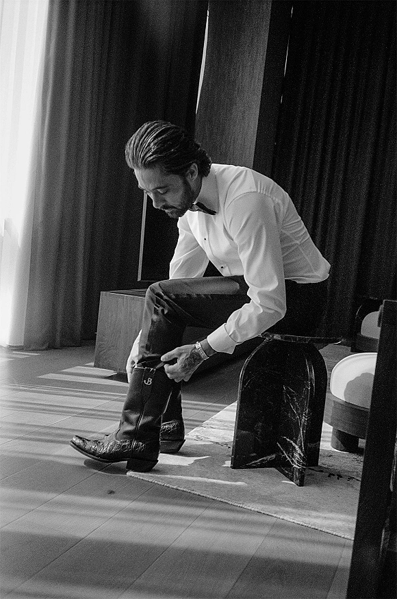 Groom getting ready, groom putting on boots in a tuxedo shirt and bow tie, adjusting cowboy boots by a bench in window light
