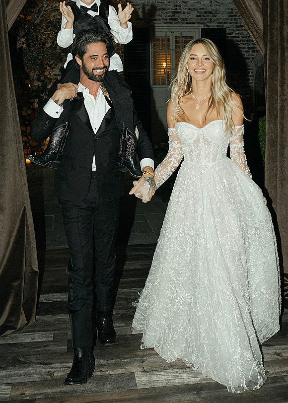 Wedding recessional as newlyweds walking out holding hands, bride in off-the-shoulder lace gown and groom in bow tie in a candlelit brick hallway