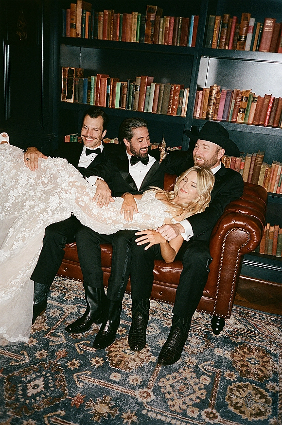 Wedding party portrait of bride with groomsmen, seated on a leather sofa in a vintage library with bookcase and rug backdrop