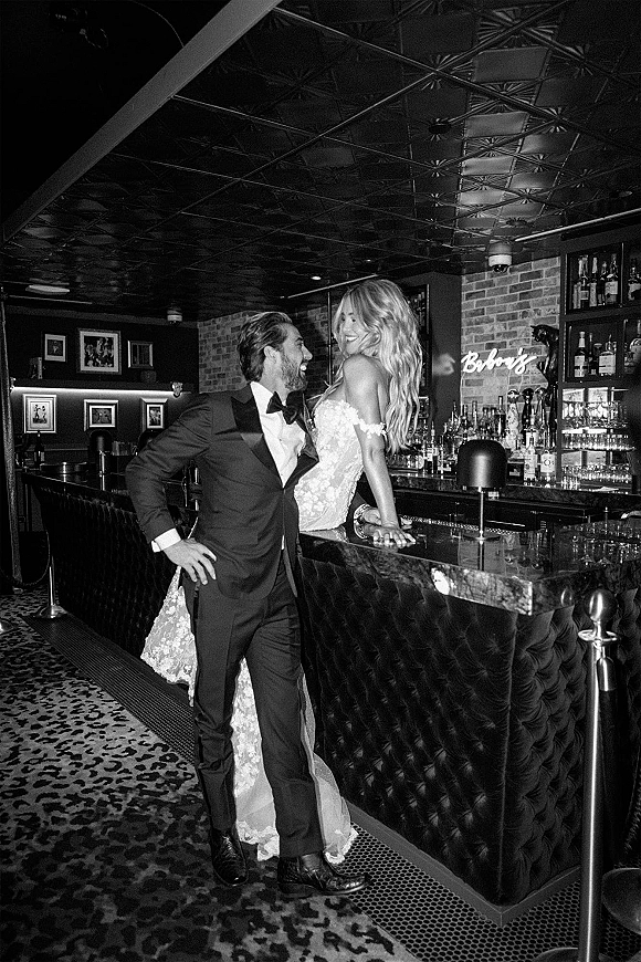 Couple portrait in a wedding bar photo, bride sitting on the tufted bar in a strapless lace dress beside groom in tuxedo, brick wall behind
